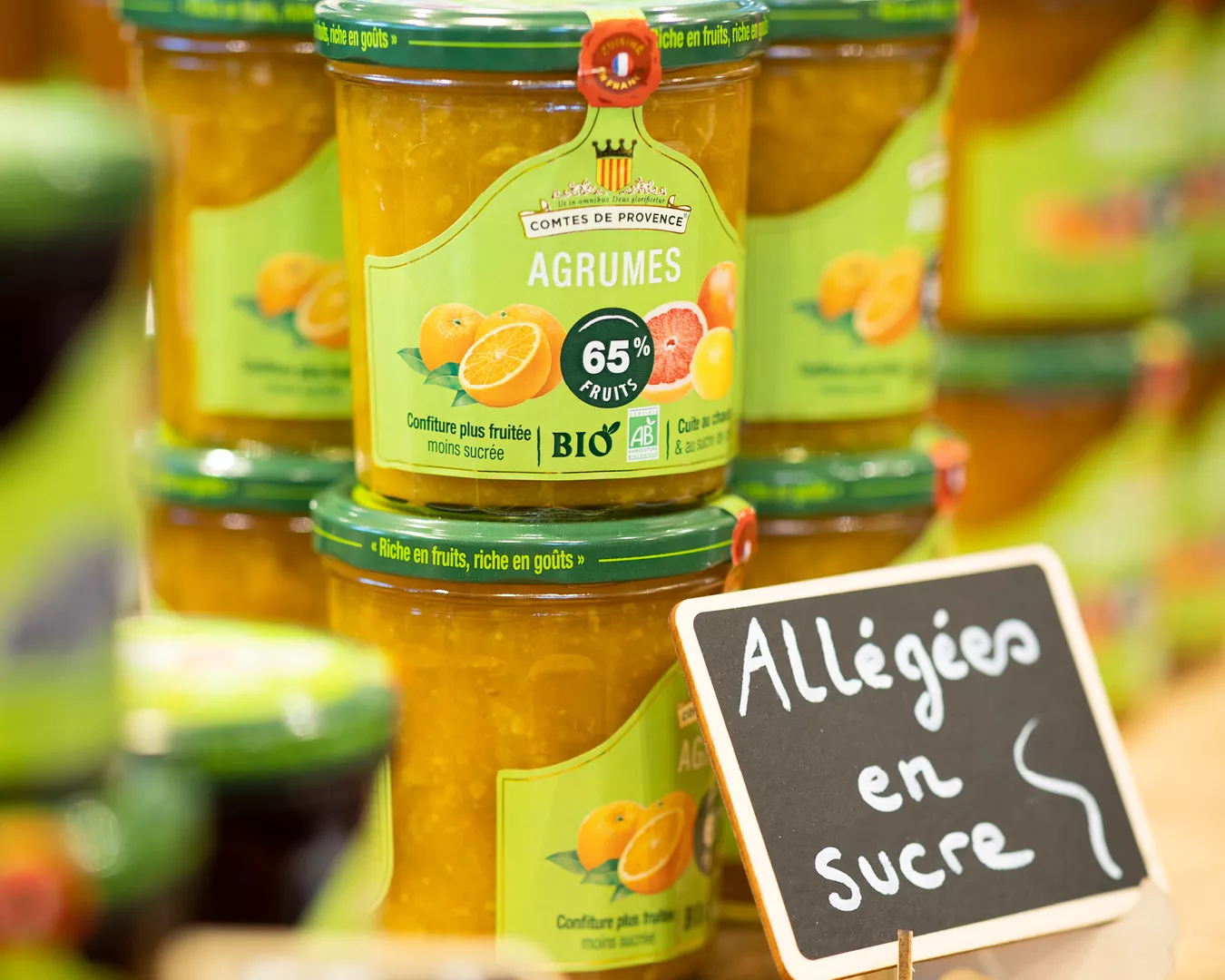 "Une série de pots de confiture biologique aux agrumes de la marque 'Comtes de Provence' est mise en avant, avec un focus sur celui de devant où figure clairement l'étiquette '65% FRUITS' et le logo BIO. En avant-plan, une ardoise avec l'inscription 'Allégée en sucre' écrit à la craie souligne la qualité diététique des produits. L'arrière-plan est flou, suggérant une abondance de ces pots de confiture sur les étagères