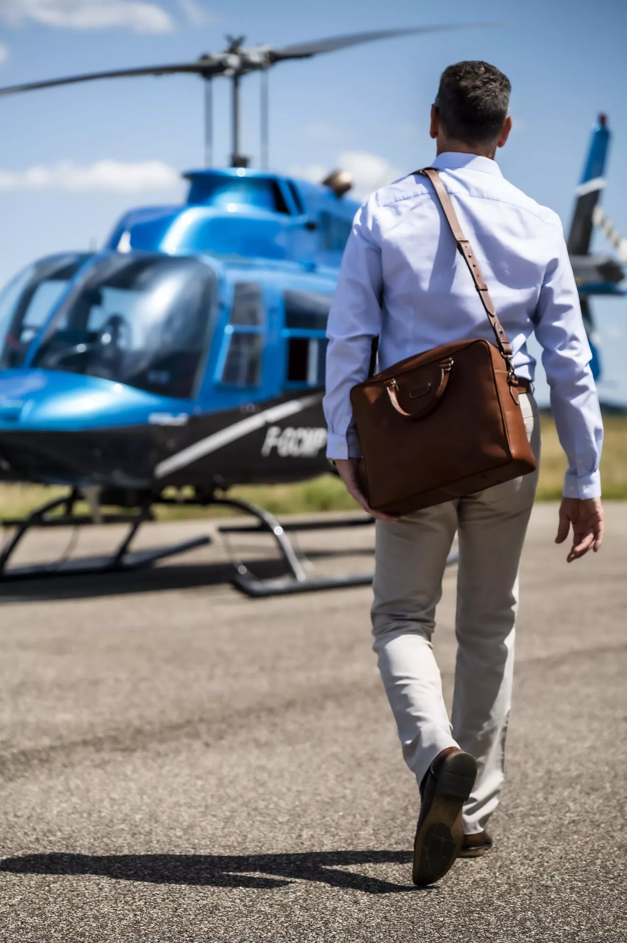 Sac 24h/ordinateur en cuir double tannage, de couleur havane, collection JAMES, porté à l'épaule du mannequin  homme qui se dirige vers un hélicoptère