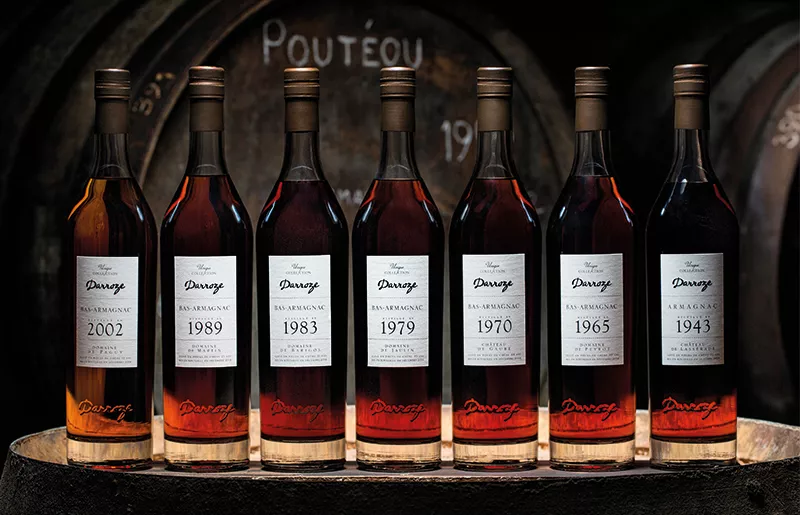 Cette image présente une série de sept bouteilles de Bas-Armagnac millésimé de la Maison Darroze, soigneusement alignées sur un tonneau en bois. Les millésimes visibles sur les étiquettes sont 2002, 1989, 1983, 1979, 1970, 1965 et 1943. Chaque bouteille arbore une étiquette blanche avec des détails noirs, indiquant le millésime et la mention "Bas-Armagnac". En arrière-plan, des barriques en bois ajoutent une touche d'authenticité et rappellent le vieillissement traditionnel de cette eau-de-vie.