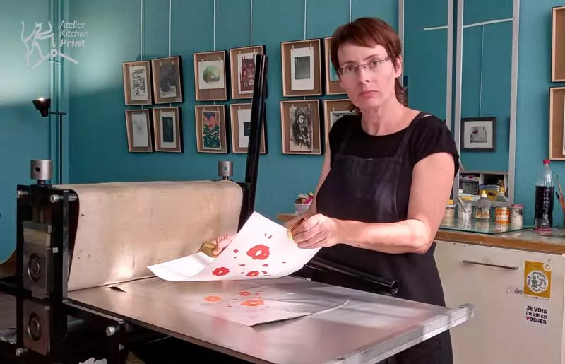 Emilie Aizier s'aprete à poser une feuille de papier sur la presse à l'aide de pinces pour ne pas salir le papier qu'elle s'aprete à imprimer. Sur le lit de la presse, il y a une feuille d'aluminium (matrice) encrée. Une fois le papier posée par-dessus la matrice, avec la pression de la presse, l'encre va imprégner le papier qui sera donc imprimé.